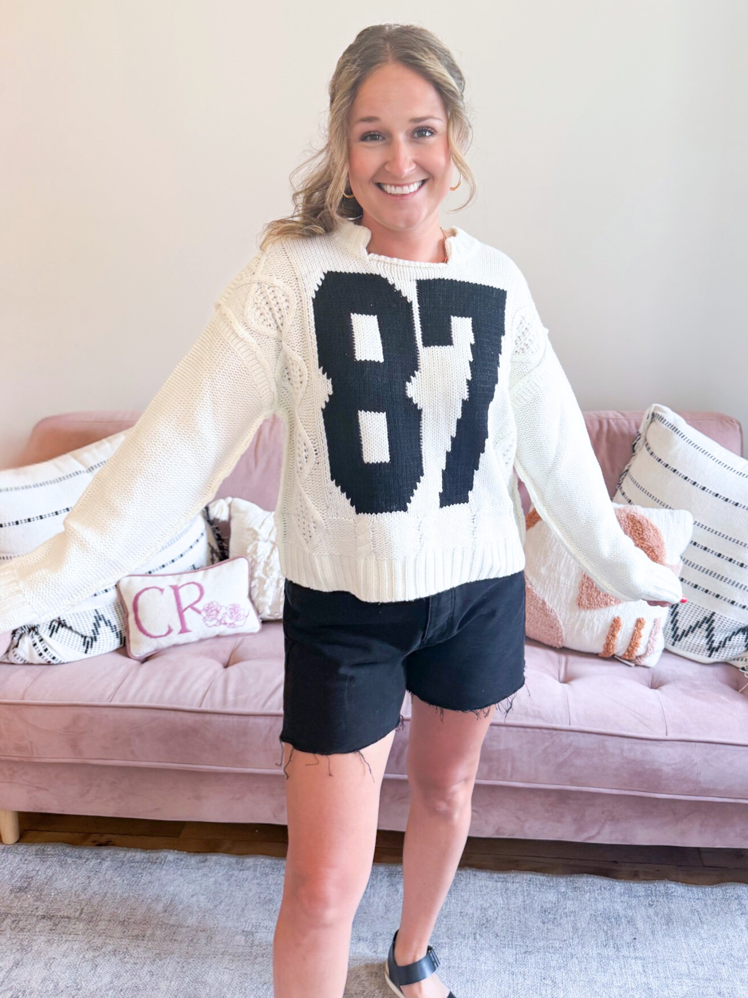87 Cable Knit Sweater. Woman wearing white Cable Knit Sweater with a bold, black number 87 on the front. Also, wearing black denim shorts