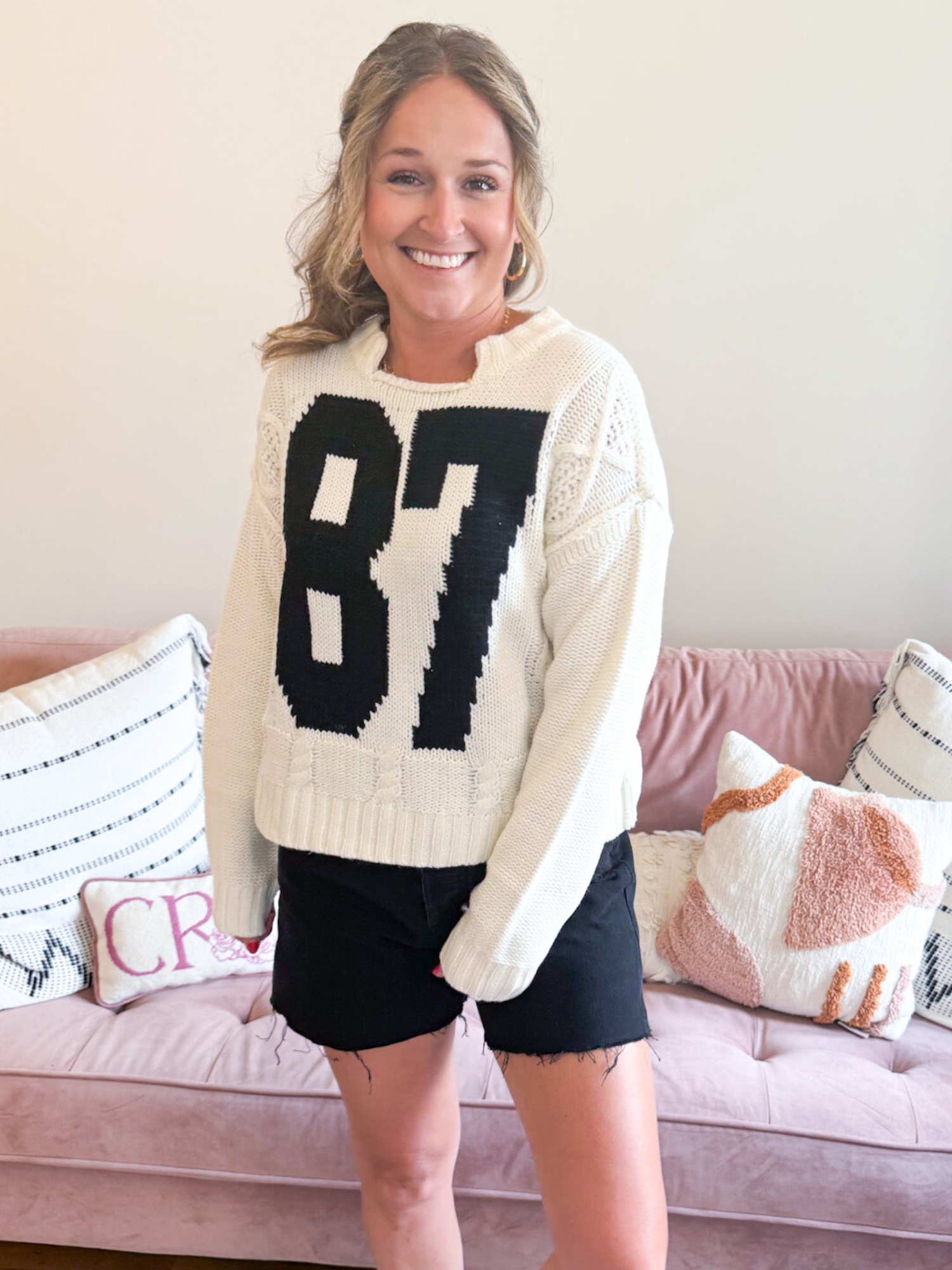 87 Cable Knit Sweater. Woman wearing white Cable Knit Sweater with a bold, black number 87 on the front. Also, wearing black denim shorts