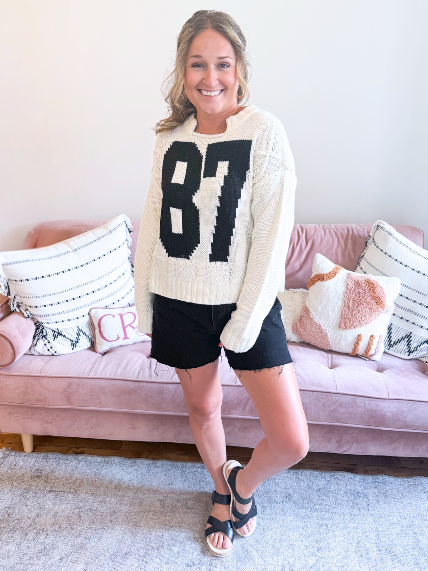87 Cable Knit Sweater. Woman wearing white Cable Knit Sweater with a bold, black number 87 on the front. Also, wearing black denim shorts