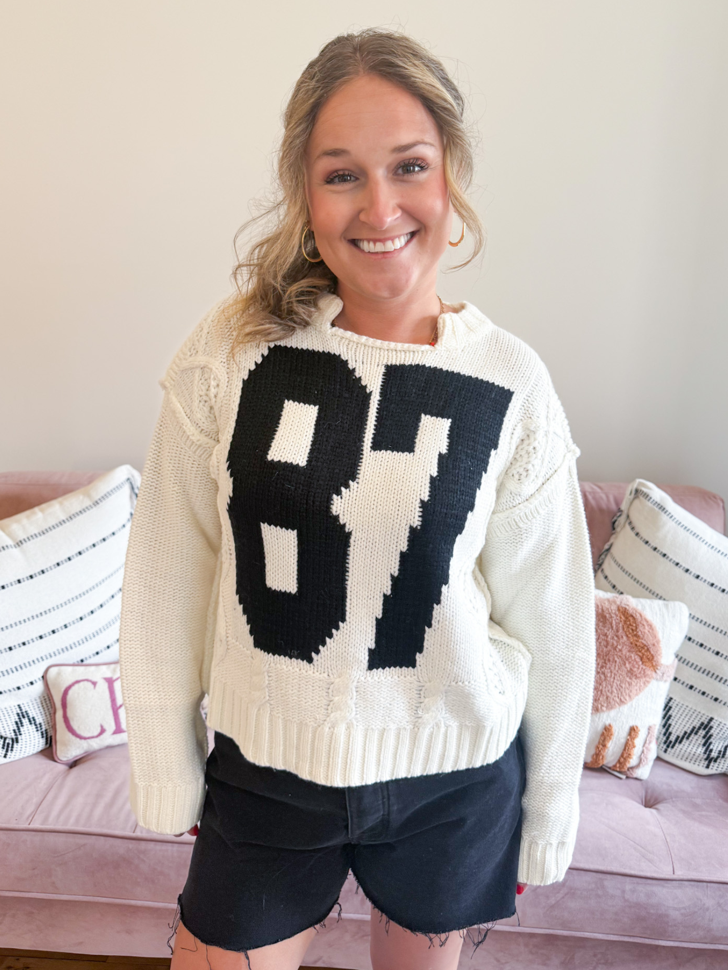87 Cable Knit Sweater. Woman wearing white Cable Knit Sweater with a bold, black number 87 on the front. Also, wearing black denim shorts